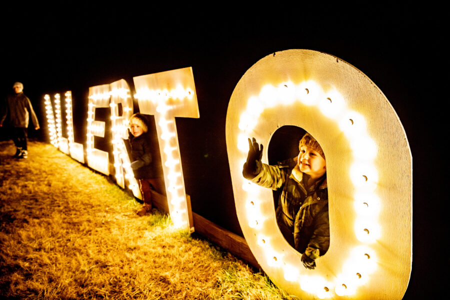Wooden letters lined with lights spelling "ALLERTON" are placed outside in the grass. Children are playing between the letters.
