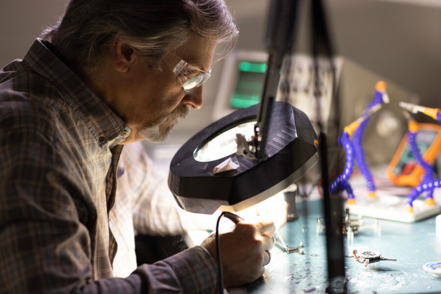 Faculty member looks through magnifying glass while soldering.