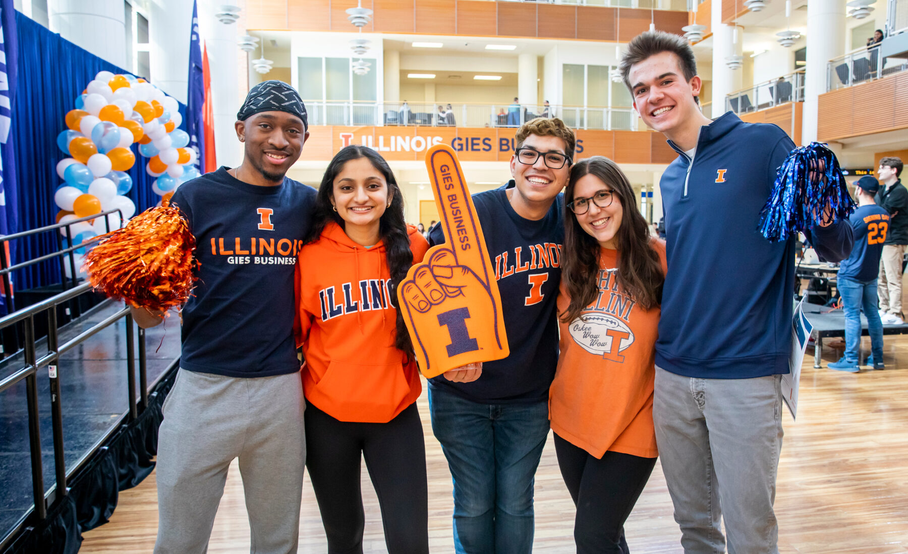 Students smiling inside of the Business building with Gies Illini Gear such as poms and foam fingers.