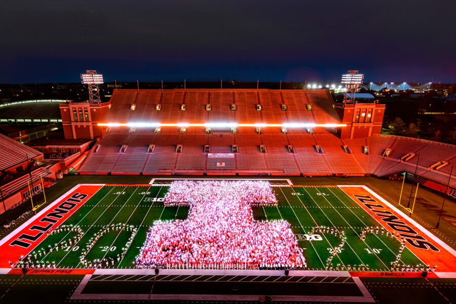 Students standing in the shape of an Illini I on the football field holding lights.