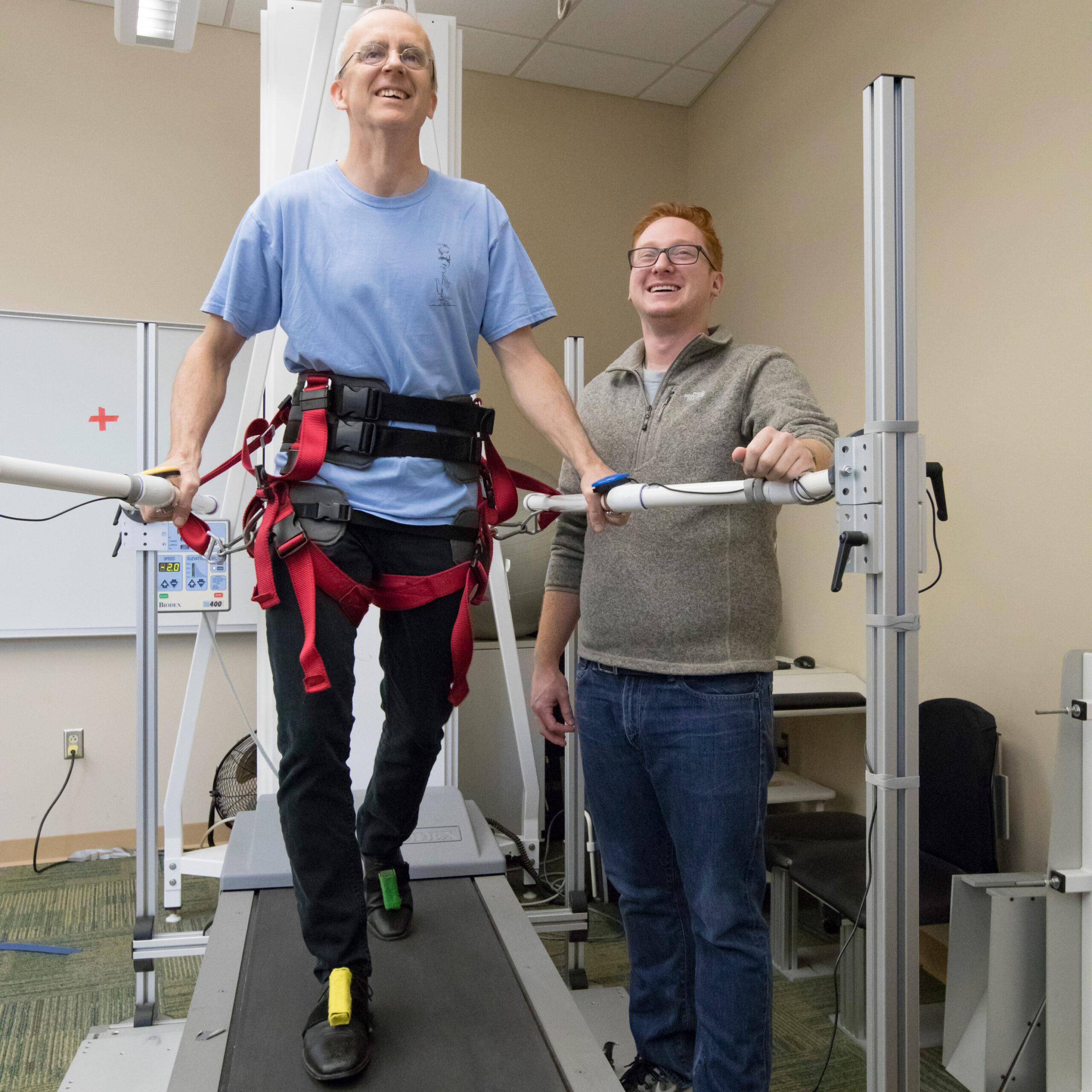A man strapped onto an elliptical smiles as he walks while another person monitors him.