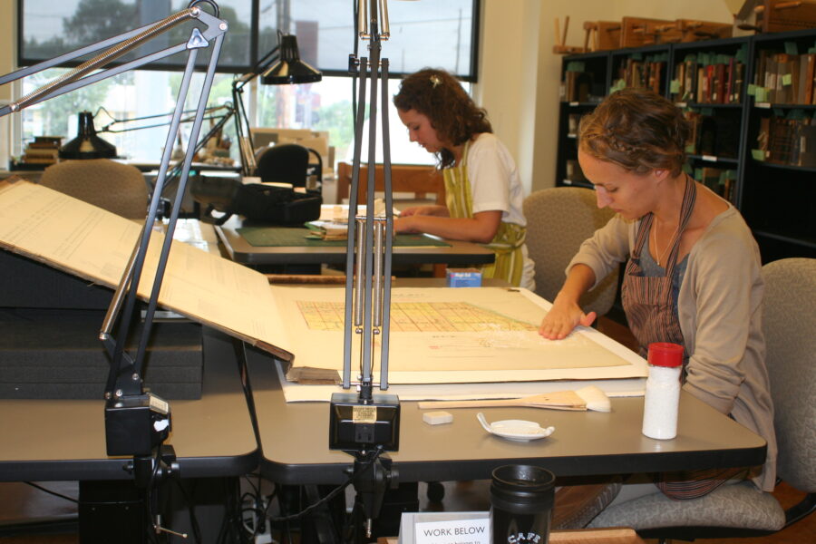 Two workers sittiing at tables with lamps as they restore rare book pages with necessary materials.