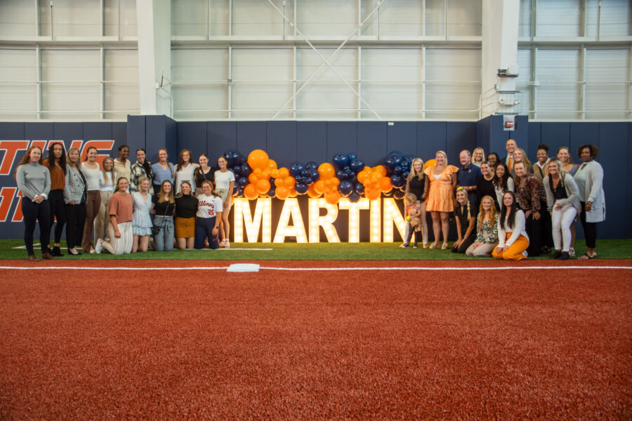 The Illinois Women's Softball team celebrates the opening of the Alice A. Martin Softball Training Center as they stand and smile together around light-up letters spelling "MARTIN" and balloons.